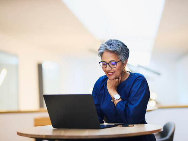 A mature woman with short gray hair and glasses smiles while working on a laptop at a round wooden table. She rests her chin on her hand in a bright, modern office setting.